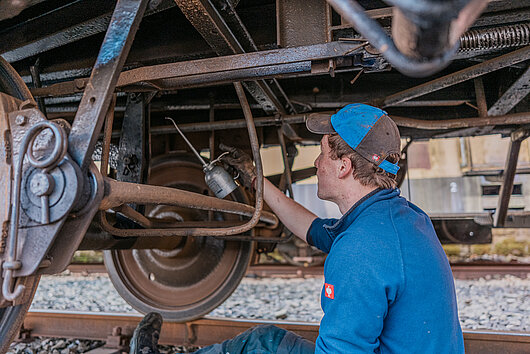 Eine Person sitzt unter einen Eisenbahnfahrzeug und hält eine Ölkanne in der Hand.